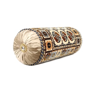 May include: A decorative cylindrical cushion with a beige satin cover and a patterned fabric insert. The fabric insert features a geometric design with brown, blue, and gold accents.