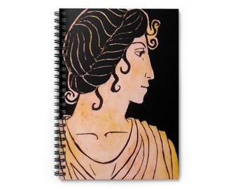 Puede incluir: Cuaderno de espiral con una ilustración clásica del perfil de una mujer. La obra de arte, en tonos negro, dorado y beige, representa a una mujer con una corona de laurel y cabello suelto sobre un fondo negro. La encuadernación en espiral del cuaderno es visible a la izquierda.