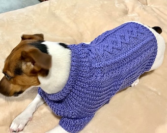 May include: A small dog wearing a purple knitted sweater with a cable knit pattern. The dog is lying on a light brown blanket.