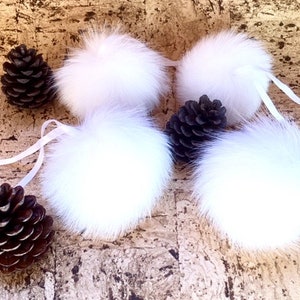 May include: Four white fluffy pom poms with white satin ribbon ties. The pom poms are arranged on a brown textured surface.