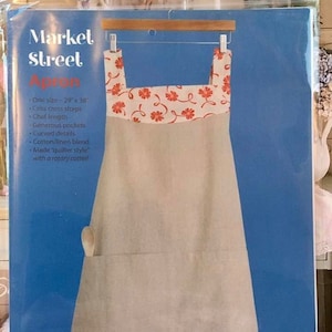 May include: A beige apron with a white and orange floral pattern on the top and bottom hem. The text on the packaging reads Market Street Apron, One size - 74 cm x 97 cm, Criss cross straps, Chef length, Generous pockets, Curved details, Cotton/linen blend.