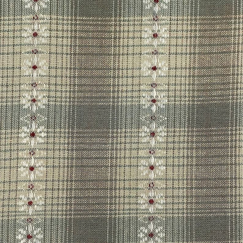 May include: Close-up of a woven fabric with a plaid pattern in shades of gray, beige, and white. Vertical rows of white floral designs with red centers are interspersed throughout the fabric. The fabric has a textured appearance.