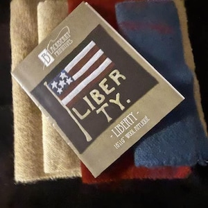 May include: A rectangular wool applique with the word "LIBERTY" and a flag design. The applique is on top of tan, red, and blue wool fabric. The tag reads "Blackberry Primitives" and "16x19 Wool Applique."