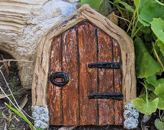 May include: A small, brown wooden door with a black metal handle and hinges. The door is surrounded by a stone frame and is set against a backdrop of green foliage and brown dirt.