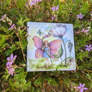 May include: A watercolor painting of a fairy with pink wings holding a flower. The fairy is wearing a blue dress and has brown hair. The painting is set against a background of green leaves and flowers.