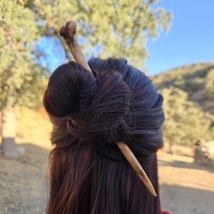 May include: A light brown, smooth, tapered wooden hair stick inserted through a dark brown hair bun. The hair accessory features a natural wood grain texture and a slightly curved, pointed end, securing the hair in a simple, functional style.
