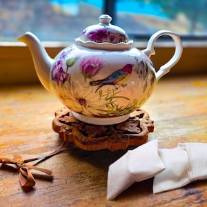 May include: A white porcelain teapot with a floral and bird design, resting on a carved wooden coaster. The teapot features pink and purple flowers and a colorful bird. Two white tea bags and a wooden stirrer are also present.