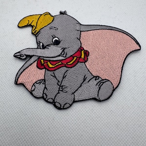 以下が含まれることがあります： 象のダンボが描かれた刺繍ワッペン。ワッペンは、ピンクの耳、黄色の帽子、赤い襟をしたグレーのダンボを描いています。このワッペンは、衣類やアクセサリーへの貼り付け用に設計されています。