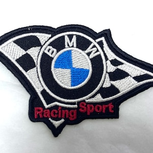Puede incluir: Bandera a cuadros blanco y negro con un logotipo BMW azul y blanco en el centro. El logotipo está rodeado del texto "Racing Sport" en rojo.