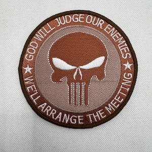 以下が含まれることがあります： スカルデザインの刺繍入り円形パッチ。パッチは茶色の縁取りで、「GOD WILL JUDGE OUR ENEMIES」と「WE'LL ARRANGE THE MEETING」の文字が特徴です。スカルは茶色で、白いアクセントが付いています。