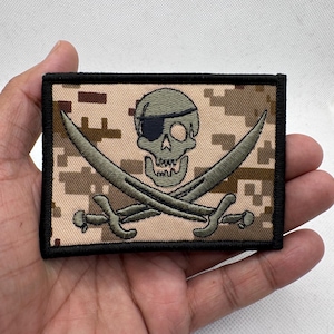 Könnte beinhalten: Rechteckiger Aufnäher mit einem Totenkopf mit Augenklappe und gekreuzten Schwertern. Der Totenkopf und die Schwerter sind grün, vor einem Tarnhintergrund in Beige und Braun, mit einem schwarzen Rand. Ideal für Jacken oder Taschen.