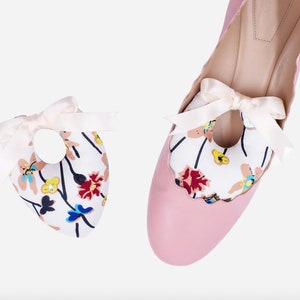 May include: A pair of pink heeled shoes with a white floral pattern on the inside. The shoes have a white satin bow on the top.