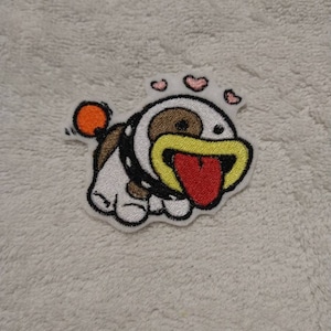 May include: An embroidered patch featuring a cartoon dog with white and brown fur, a black collar, a yellow mouth, and a large red tongue sticking out. The design includes a small orange ball on a stick and three small pink hearts floating above the head.