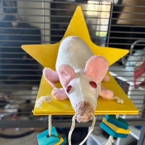 May include: A white plush rat toy with pink ears and red eyes rests on a yellow star-shaped platform. The platform is part of a hanging toy with colorful wooden blocks and string, designed for small pets.