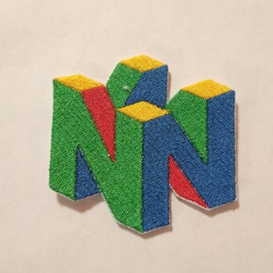 May include: An embroidered patch featuring a stylized letter N design. The geometric shape is composed of vibrant green, blue, red, and yellow thread. The design has a three-dimensional appearance with distinct colored segments and a white border.