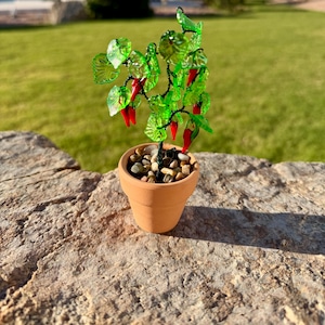 May include: A miniature decorative plant featuring translucent green glass leaves and small red glass chili peppers attached to dark wire branches. The plant is potted in a small, light brown terracotta pot filled with smooth, multi-colored pebbles.