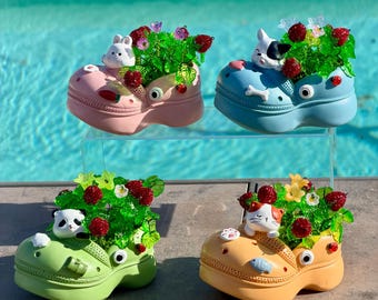 May include: Four platform clogs in pink, blue, light green, and orange, each featuring a small animal figurine, green foliage, and red berry accents. The clogs are decorated with small charms including a carrot, bone, bamboo, and fish.