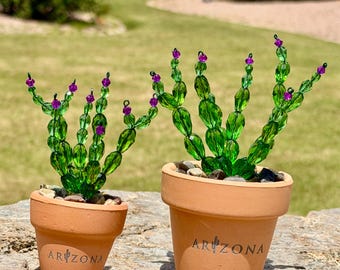 May include: Two decorative cactus sculptures in terracotta pots. The cacti are made of green glass beads with purple accents, and the pots are inscribed with "ARIZONA". The pots are filled with small stones.