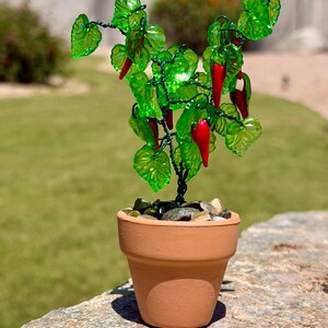May include: A miniature decorative plant featuring translucent green glass leaves and small red glass chili peppers attached to a dark wire frame. The plant is potted in a small terracotta-colored clay pot filled with smooth, multi-colored pebbles.