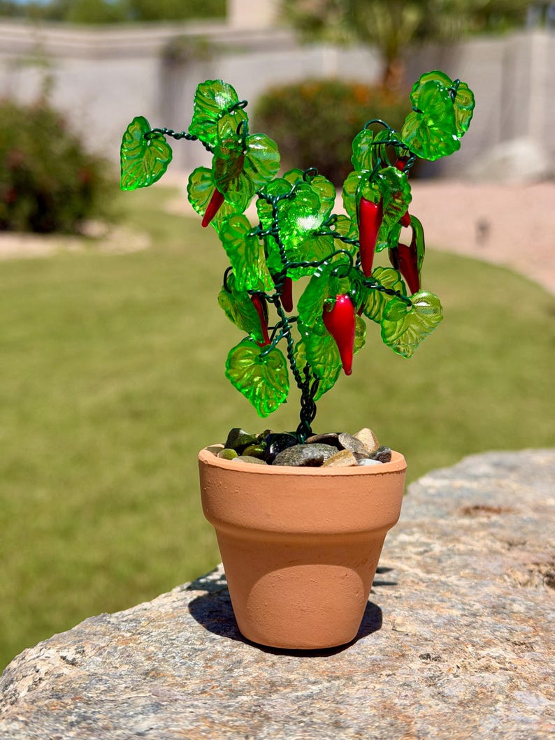 May include: A miniature decorative plant featuring translucent green glass leaves and small red glass chili peppers attached to a dark wire frame. The plant is potted in a small terracotta-colored clay pot filled with smooth, multi-colored pebbles.