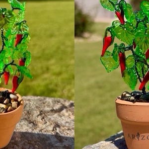 May include: A decorative plant sculpture featuring bright red glass chili peppers and translucent green glass leaves on dark wire stems, potted in a small terracotta clay pot filled with smooth river stones, with the word ARIZONA printed on the front.