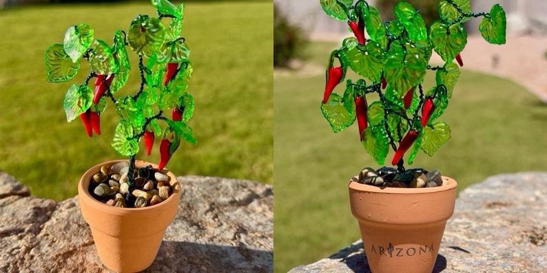May include: A decorative plant sculpture featuring bright red glass chili peppers and translucent green glass leaves on dark wire stems, potted in a small terracotta clay pot filled with smooth river stones, with the word ARIZONA printed on the front.