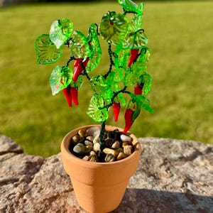 May include: A decorative miniature plant sculpture featuring bright red glass chili peppers and translucent green glass leaves attached to dark wire branches. The plant is potted in a small terracotta-colored clay pot filled with smooth pebbles.