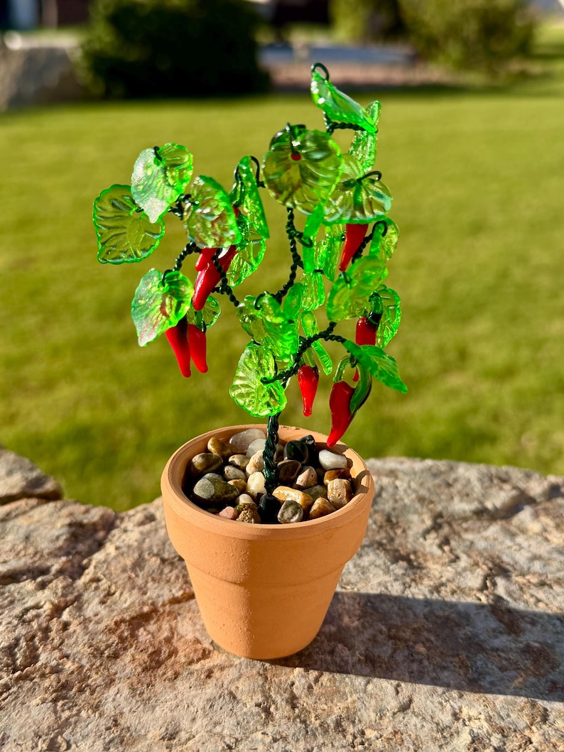 May include: A decorative miniature plant sculpture featuring bright red glass chili peppers and translucent green glass leaves attached to dark wire branches. The plant is potted in a small terracotta-colored clay pot filled with smooth pebbles.