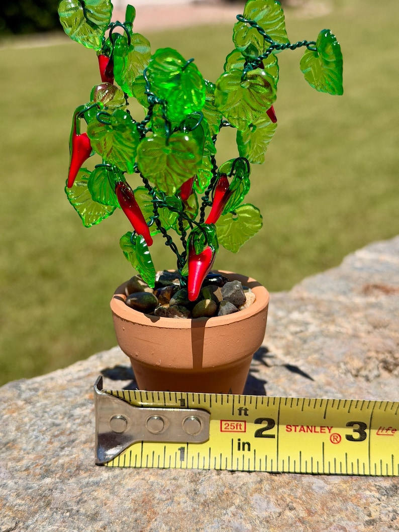 May include: A miniature glass chili pepper plant with vibrant green leaves and bright red peppers, set in a small terracotta pot filled with dark pebbles. A yellow Stanley measuring tape shows the pot has a diameter of approximately 2 inches.