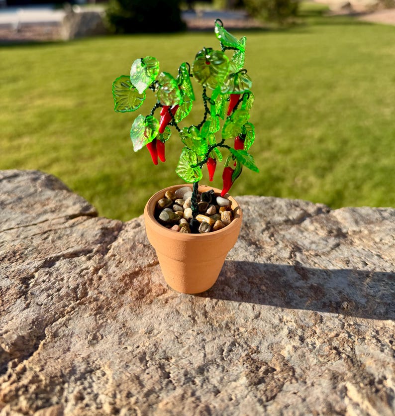 May include: A miniature decorative plant featuring translucent green glass leaves and small red glass chili peppers attached to dark wire branches. The plant is potted in a small, light brown terracotta pot filled with smooth, multi-colored pebbles.