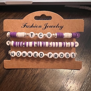 May include: Three purple and white beaded bracelets with the words "TCU" and "Horned Frogs" spelled out in black letters on white beads.