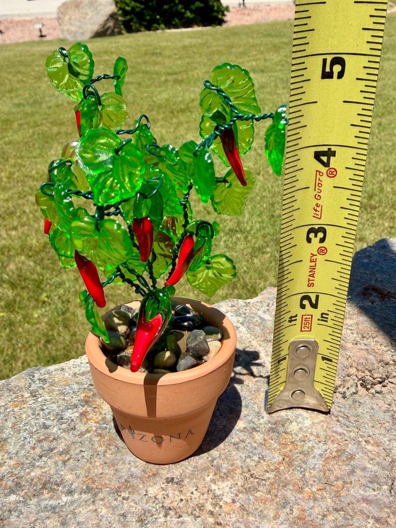 May include: A decorative glass chili pepper plant with bright red peppers and translucent green leaves on wire stems, potted in a small terracotta pot filled with smooth pebbles. A yellow measuring tape shows the plant is 5 inches tall.