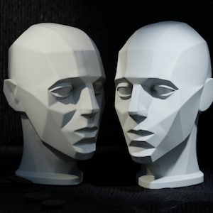 May include: Two white plaster busts of human heads, each with a geometric, faceted design. The busts are facing each other, with the left bust slightly tilted to the right.