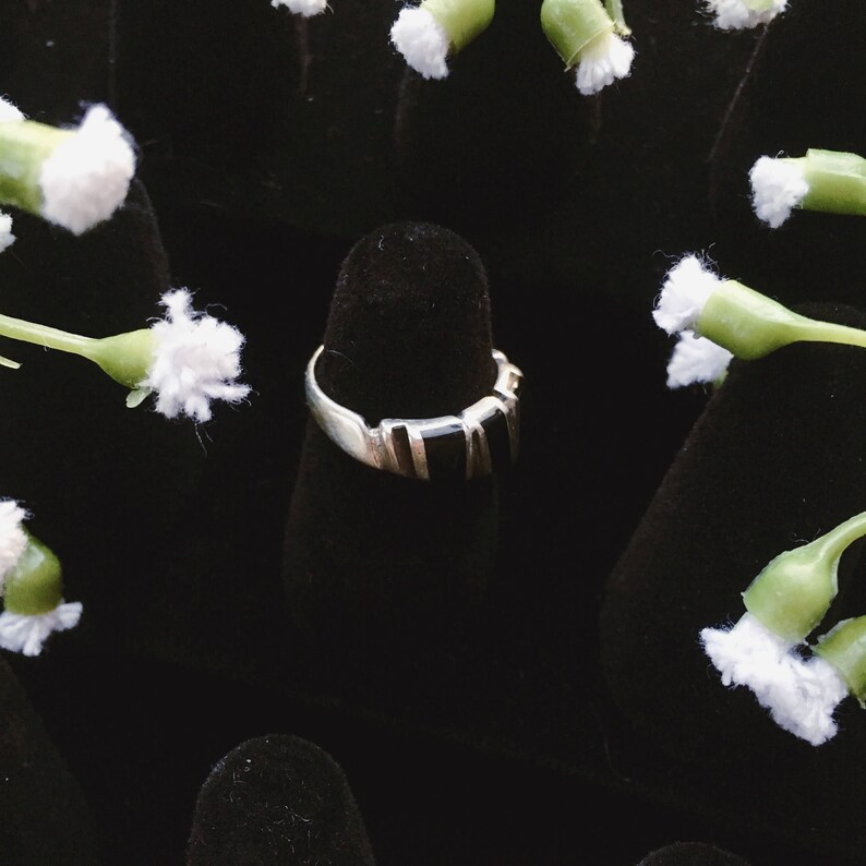 May include: A silver ring with a black stone set in a rectangular shape. The ring is displayed on a black velvet ring holder.