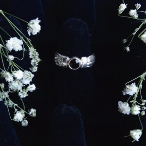 May include: A silver ring with a dark red gemstone set in the center. The ring has a textured design on the band and is displayed on a black surface, with white flowers in the background.