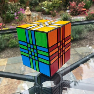 Könnte beinhalten: Ein mehrfarbiger Würfelpuzzle mit einem schwarzen Gittermuster auf jeder Seite. Der Würfel besteht aus kleineren Würfeln, die jeweils eine andere Farbe haben. Die Farben sind Gelb, Orange, Rot, Grün, Blau und Weiß.