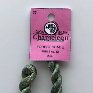 May include: A skein of green and blue Chameleon Forest Shade embroidery thread, Perle no. 12, 20m, is displayed. The thread is attached to a pink tag with the brand logo and product information. The tag reads "Hand Coloured Embroidery Thread."