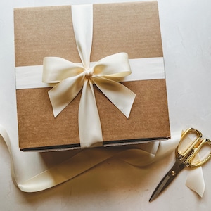 May include: A brown cardboard box with a white satin ribbon tied in a bow. The ribbon is tied in a knot in the center of the box. A pair of gold scissors are lying on a white surface next to the box.