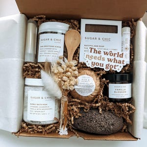 May include: A gift box filled with bath and body products, including a natural bar soap, bath soak, body scrub, bath bomb, and a loofah. The box is decorated with dried flowers and a sticker that says "The world needs what you got".