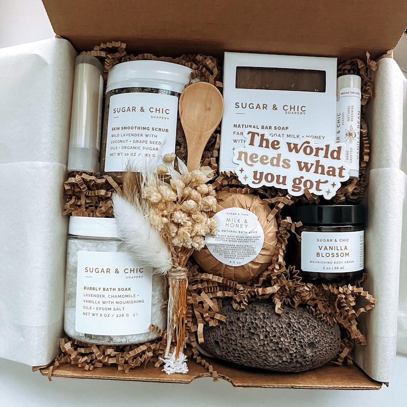 May include: A gift box filled with bath and body products, including a natural bar soap, bath soak, body scrub, bath bomb, and a loofah. The box is decorated with dried flowers and a sticker that says "The world needs what you got".