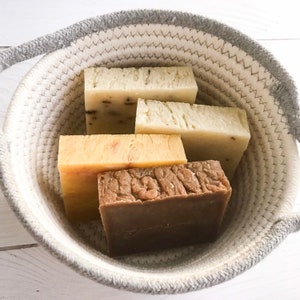 May include: A woven basket with a gray rim holds five rectangular bars of soap. The soaps vary in color, including light beige, yellow, and dark brown. Some soaps have visible textures and embedded elements.
