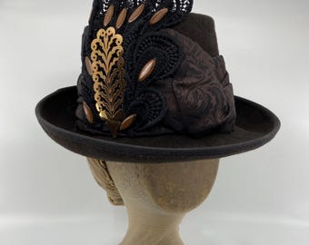 May include: A dark brown felt hat featuring a wide brim and a structured crown. The hat is adorned with a black lace applique, a metallic gold-toned feather-shaped embellishment, and several smooth, oval-shaped brown beads arranged in a fan pattern.