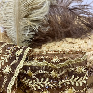 May include: A brown and gold embroidered trim with a delicate lace pattern and small brown beads. The trim is on a straw hat with brown and beige feathers.