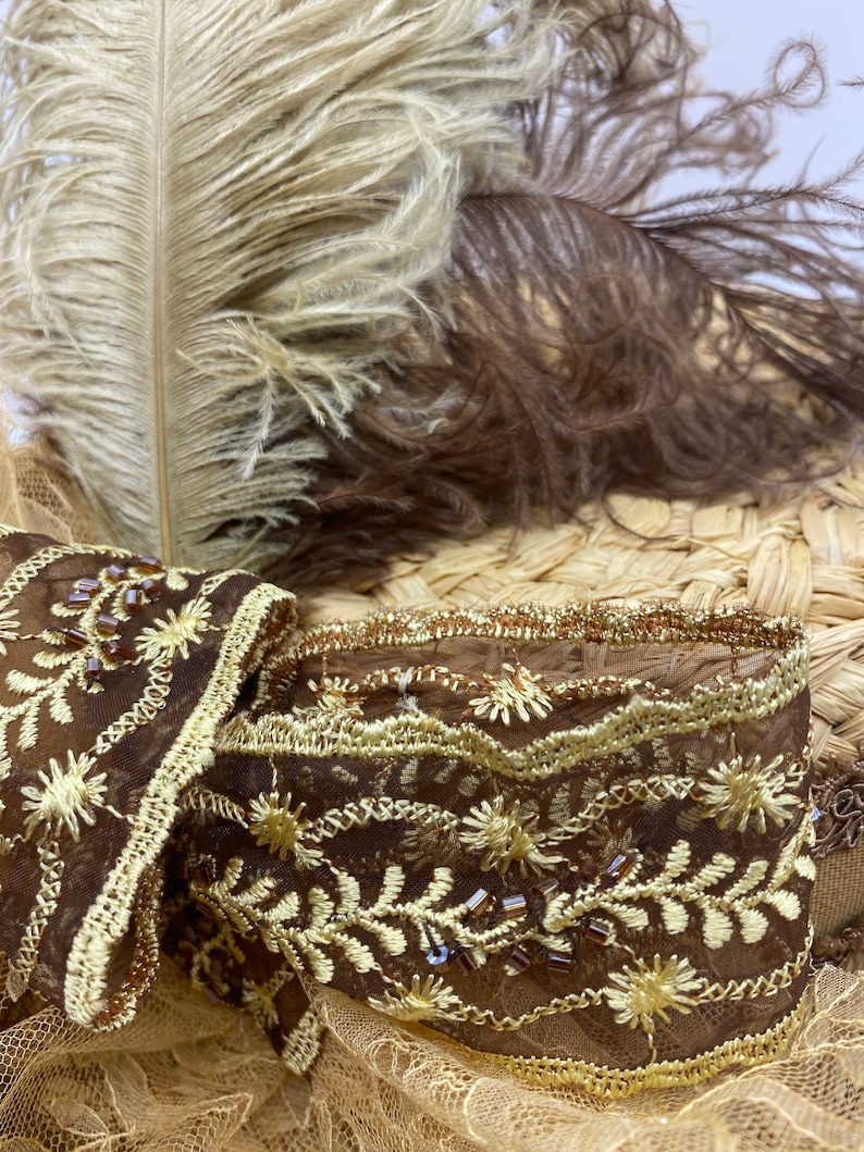 May include: A brown and gold embroidered trim with a delicate lace pattern and small brown beads. The trim is on a straw hat with brown and beige feathers.