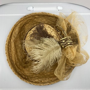May include: A brown straw hat with a brown and white feather and a brown and gold ribbon. The hat is decorated with a brown and gold tulle bow.