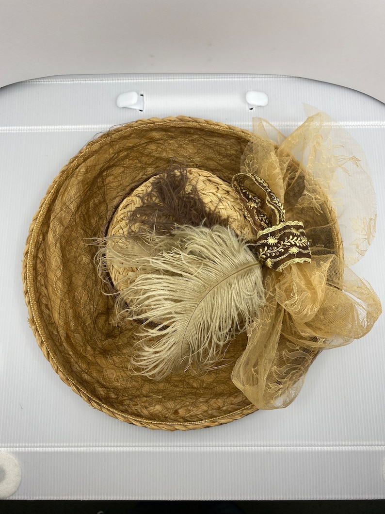 May include: A brown straw hat with a brown and white feather and a brown and gold ribbon. The hat is decorated with a brown and gold tulle bow.