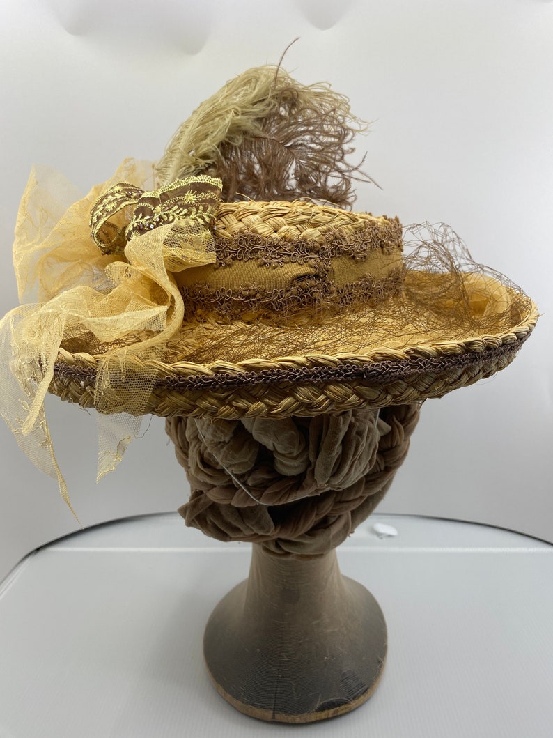 May include: A straw hat with a brown and yellow ribbon and a brown feather. The hat is decorated with lace and has a yellow veil.