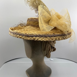 May include: A wide-brimmed straw hat with a brown braided trim and a large bow of beige netting. A single feather is attached to the top of the hat.