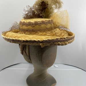 May include: A straw hat with a wide brim and a brown ribbon. The hat is decorated with a brown feather and a sheer veil. The hat is sitting on a wooden hat form.