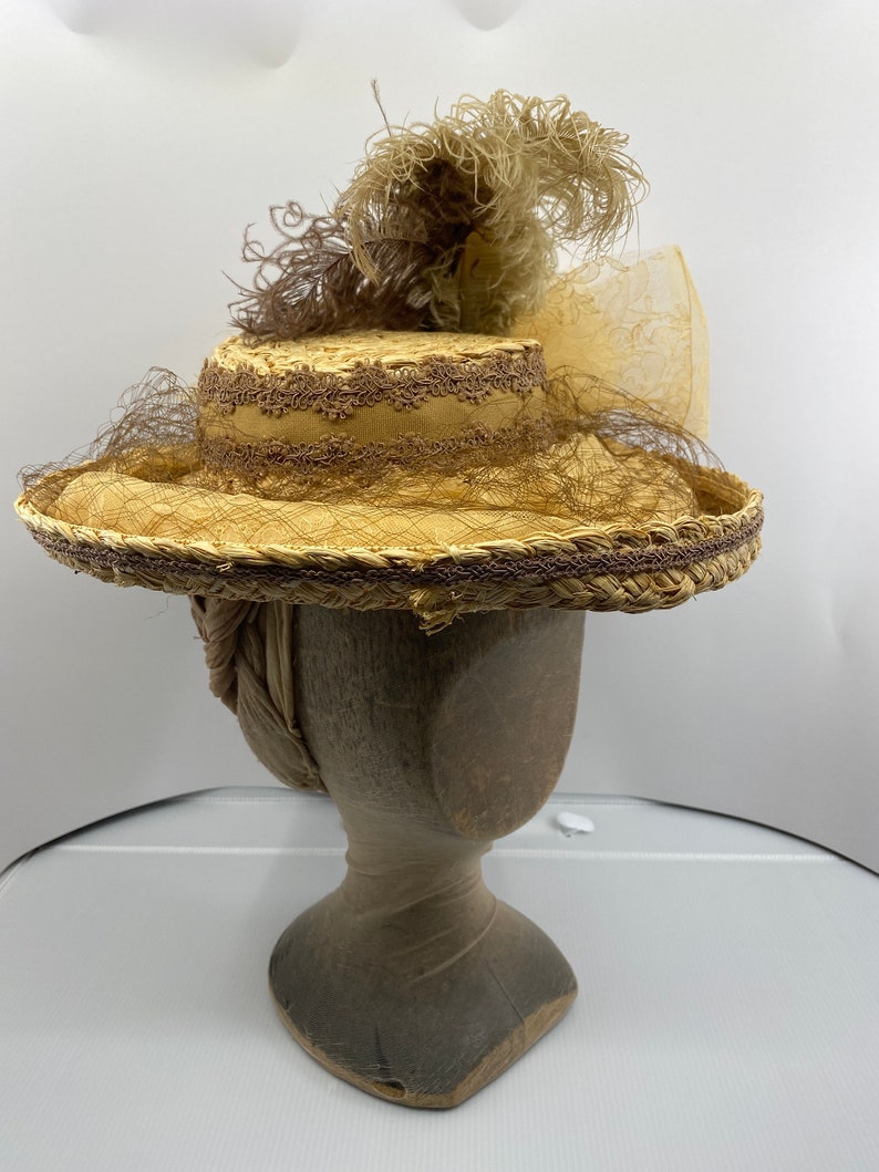 May include: A straw hat with a wide brim and a brown ribbon. The hat is decorated with a brown feather and a sheer veil. The hat is sitting on a wooden hat form.